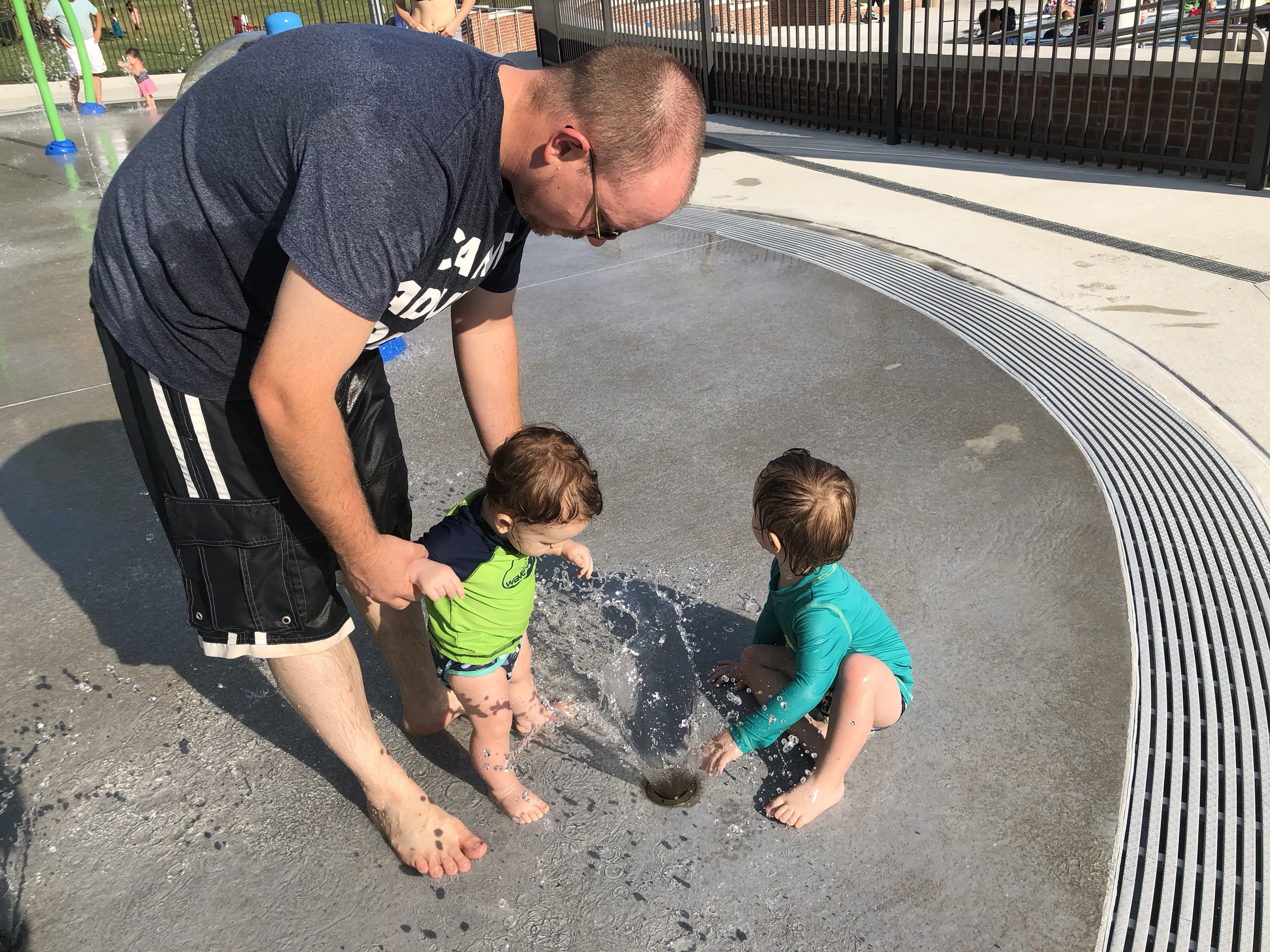 Daddy helping Shiloh stand to explore the sprinkler and Asher sitting and looking into the distance