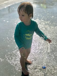 Asher in a rash guard yelling YAY while checking out the splash pad sprinkler