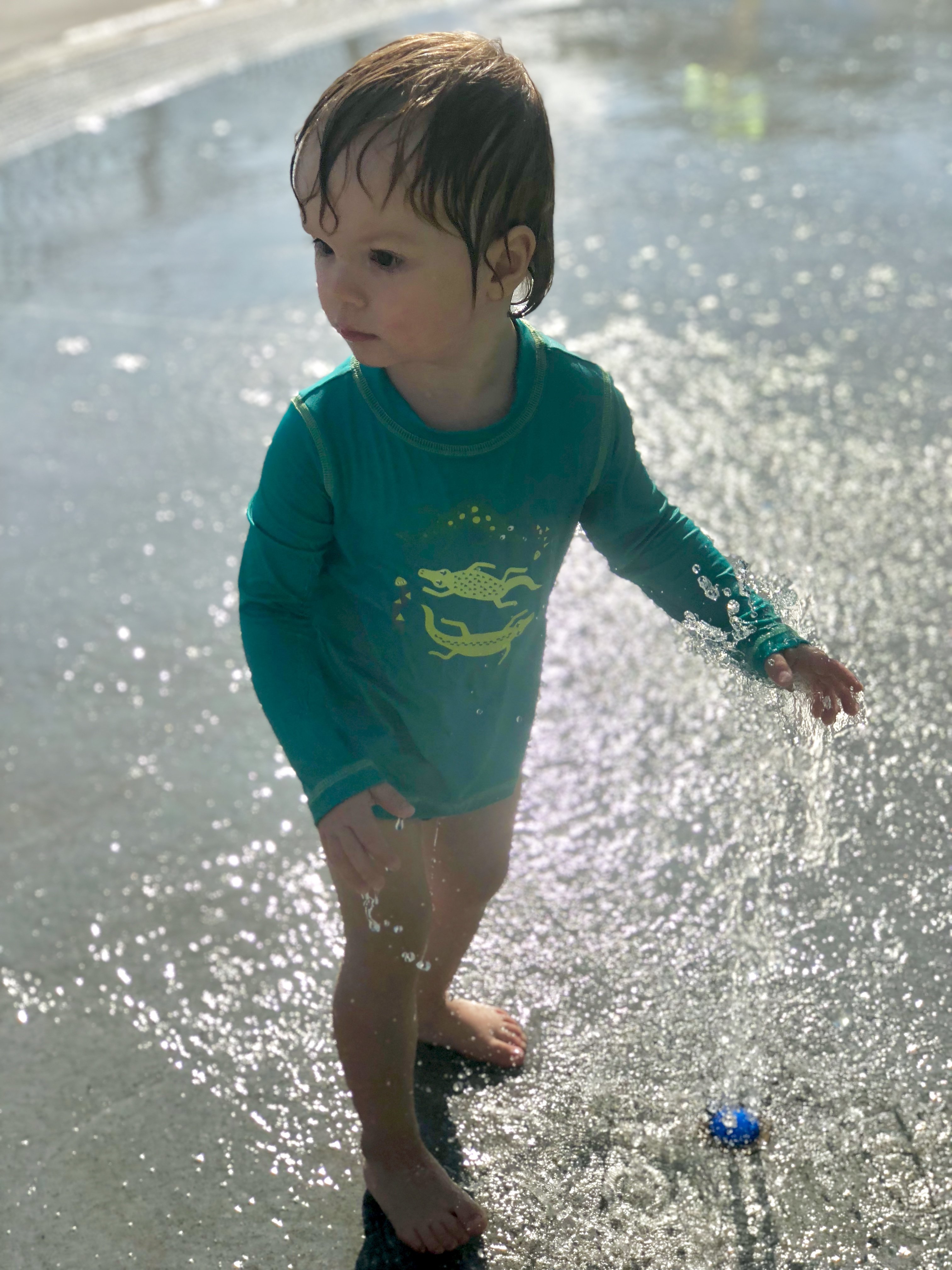 Asher in a rash guard yelling YAY while checking out the splash pad sprinkler