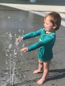 Asher in a rash guard yelling YAY while checking out the splash pad sprinkler