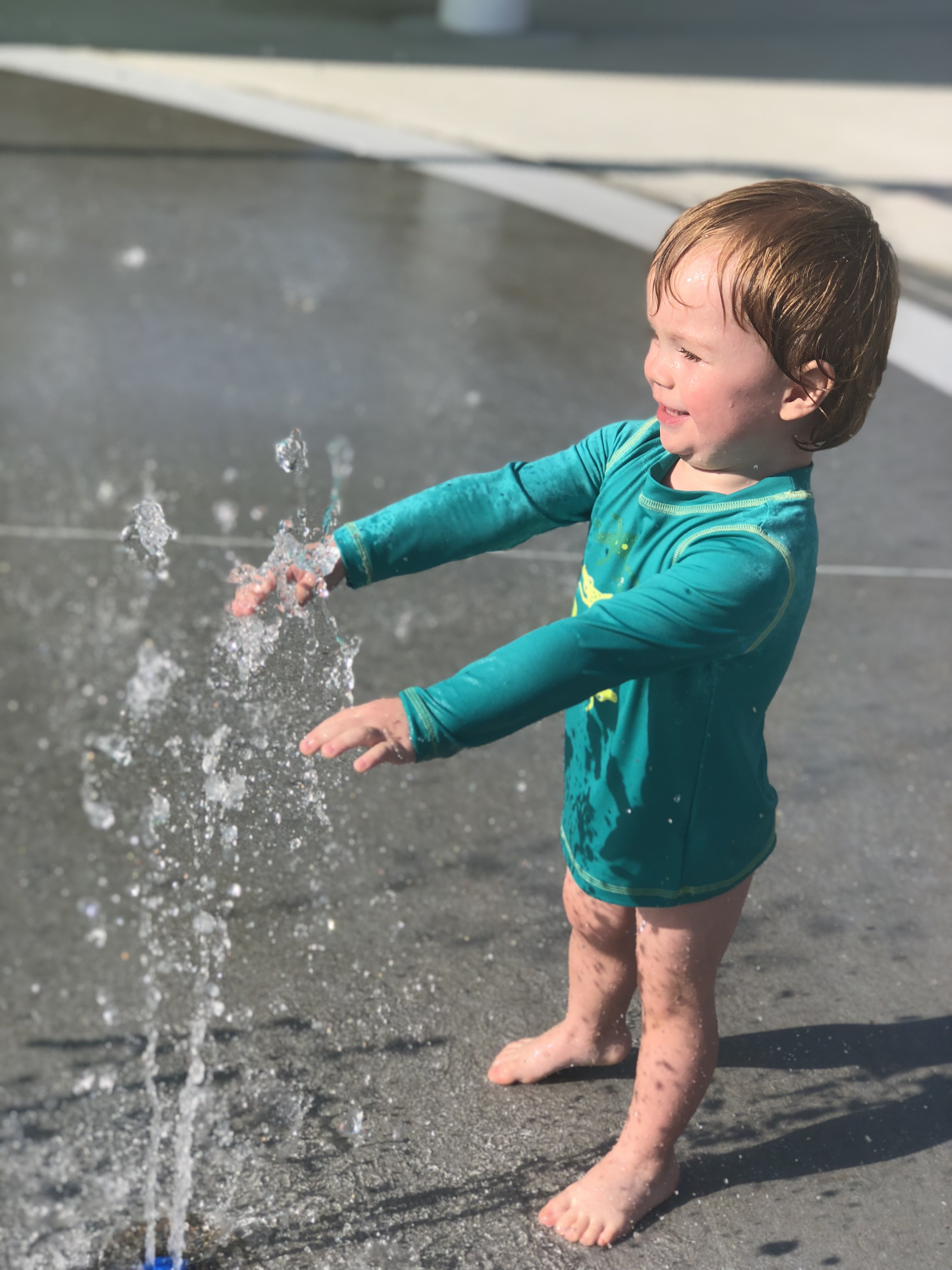 Asher in a rash guard yelling YAY while checking out the splash pad sprinkler