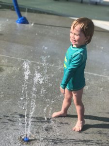 Asher in a rash guard yelling YAY while checking out the splash pad sprinkler
