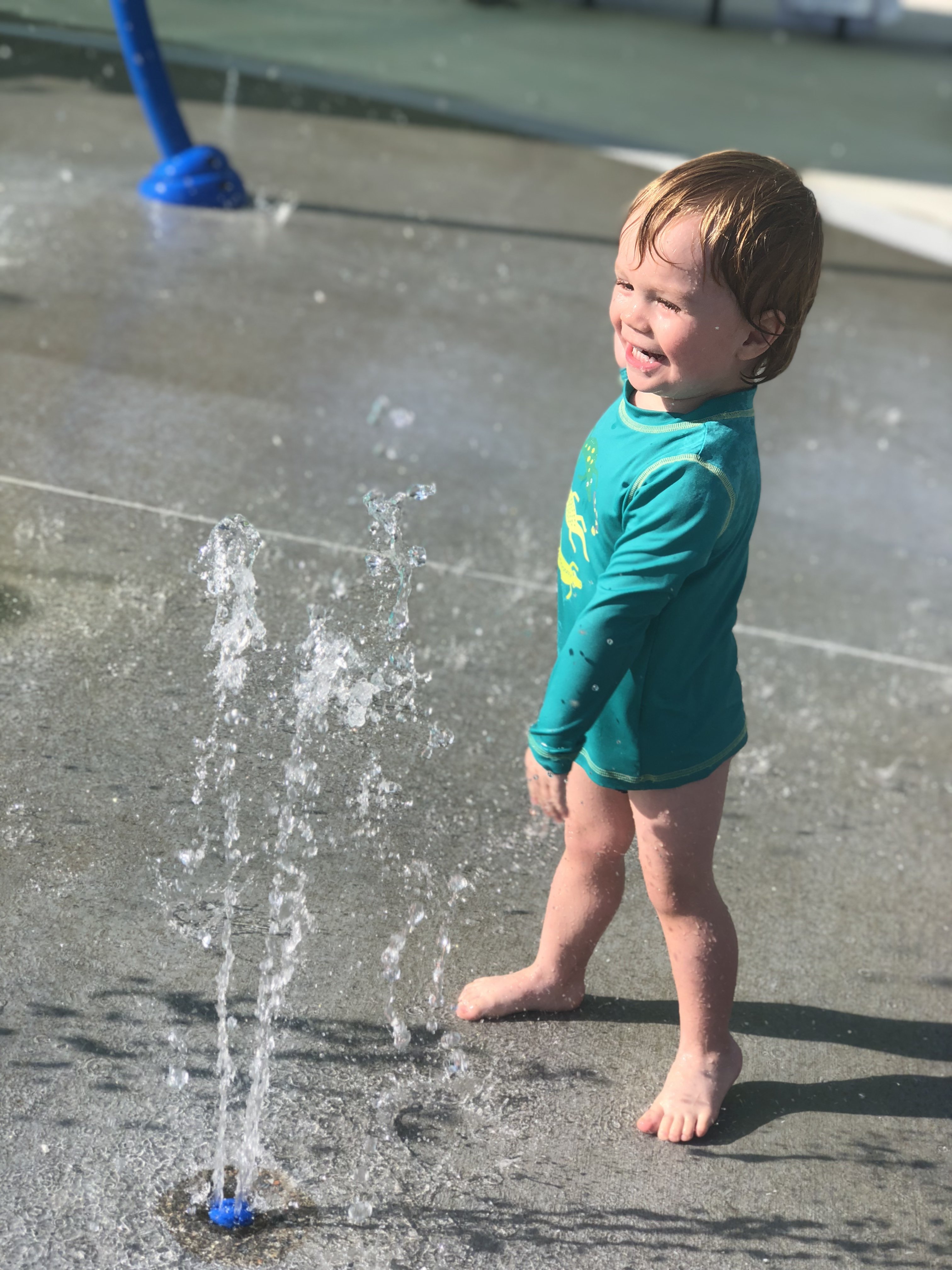 Asher in a rash guard yelling YAY while checking out the splash pad sprinkler
