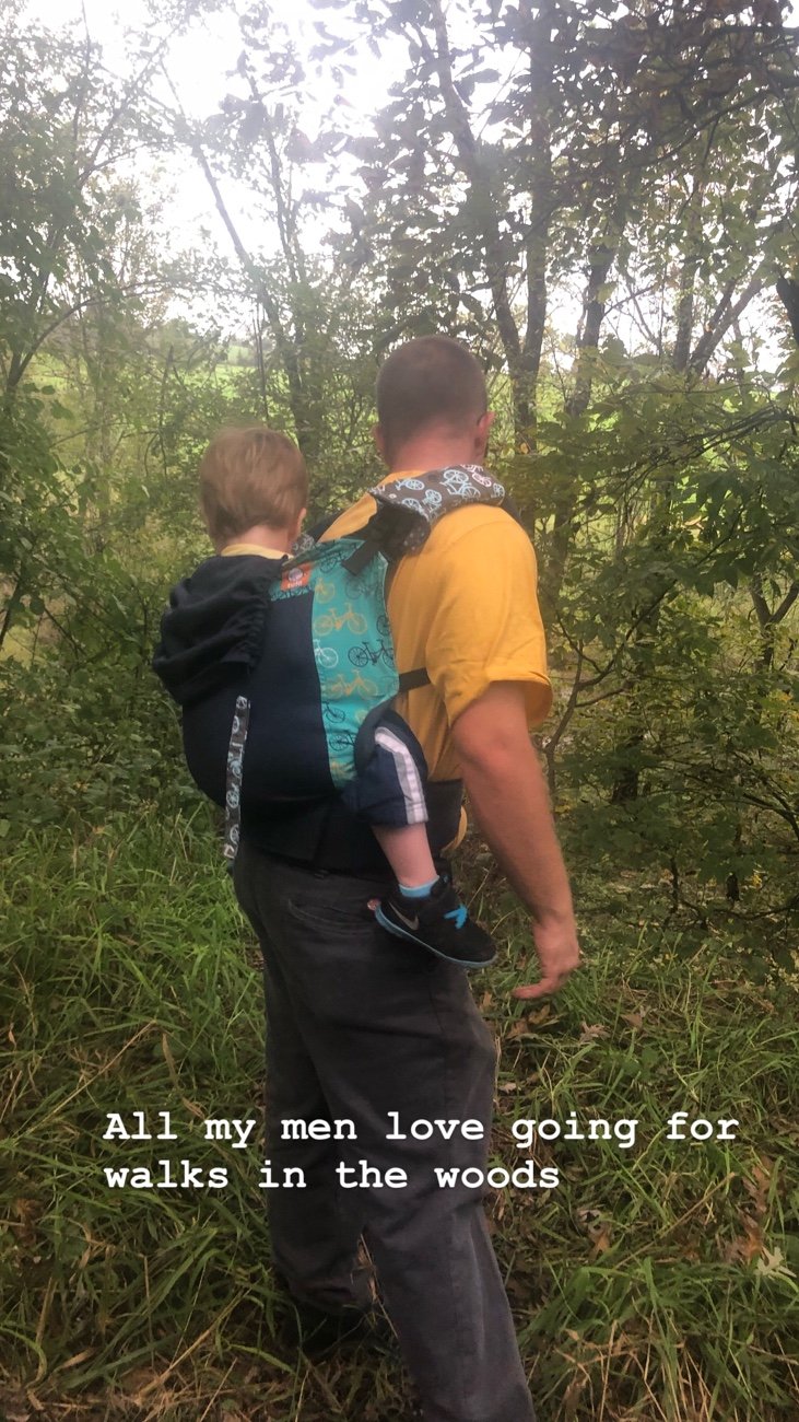 Asher on Daddy's back while walking on a grass abandoned road.  Asher is in a Tula baby carrier overlooking a stream .