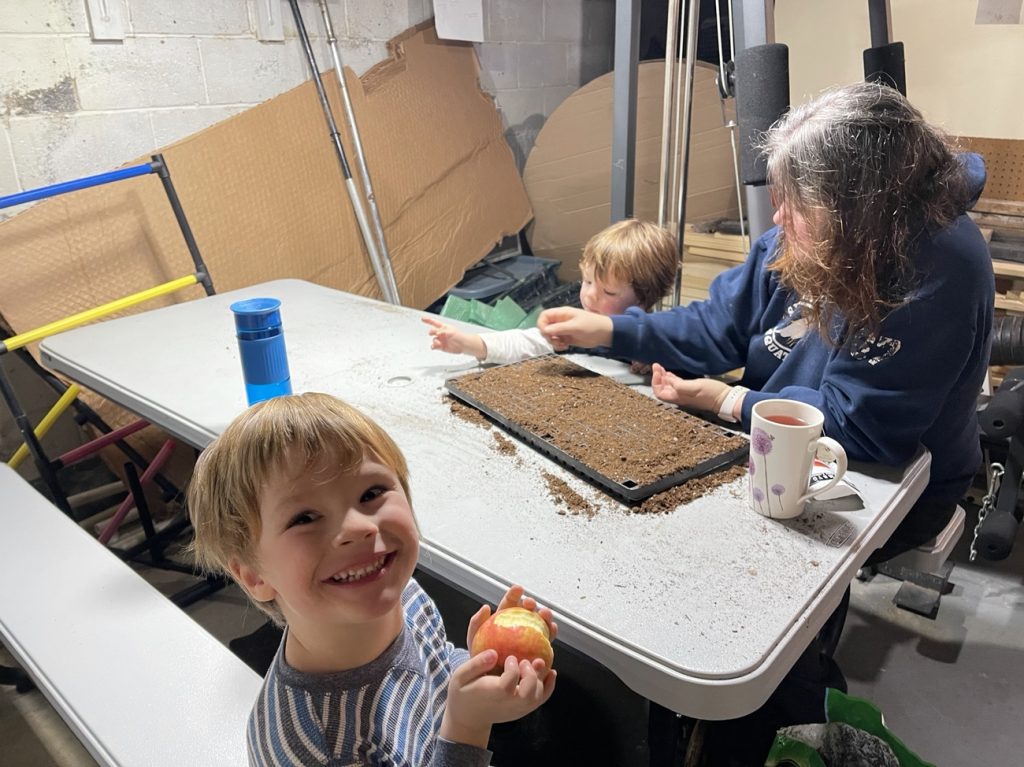 4 year old blond boy smiling snacking on an apple while mom helps 3 year old plant seeds